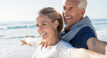 Happy senior couple standing on beach with arms outstretched and looking away. Happy couple at beach on a bright sunny day. Retired husband and smiling wife thinking about their future.