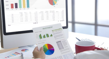 Businessman in front of modern computer holding a red coffee cup looking into investment document with colorful pie graph for investment risk analysis